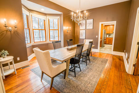 A cozy dining room with a wooden table, black chairs, and a chandelier under warm brown walls and bay windows.