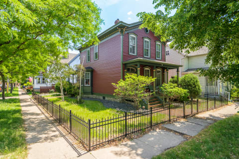 Victorian home with burgundy siding and green trim, surrounded by greenery and a wrought iron fence.
