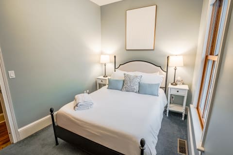 A cozy bedroom featuring a neatly made bed with white linens and blue accents, illuminated by warm bedside lamps.