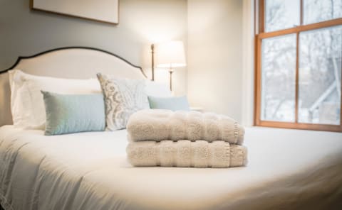 A serene bedroom with a neatly made bed, soft pillows, and stacked white towels, bathed in natural light.