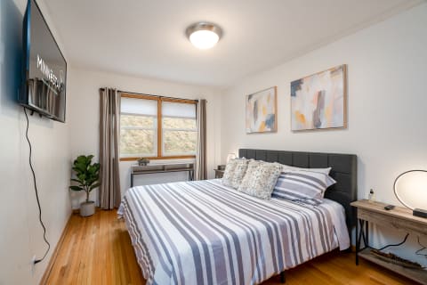 A cozy bedroom with a striped bed, wooden decor, and abstract wall art, illuminated by natural light.