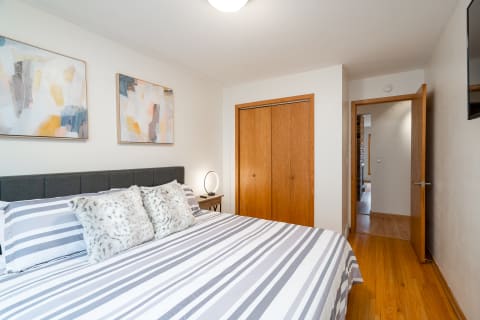 A serene bedroom featuring a comfortable bed with striped linens and faux fur pillows, abstract artwork, and wooden accents.