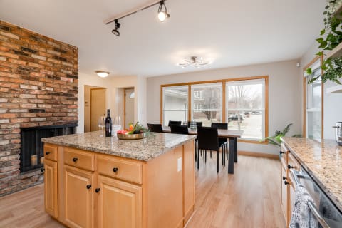 Modern kitchen with a granite island, rustic brick wall, and a dining area with large windows.