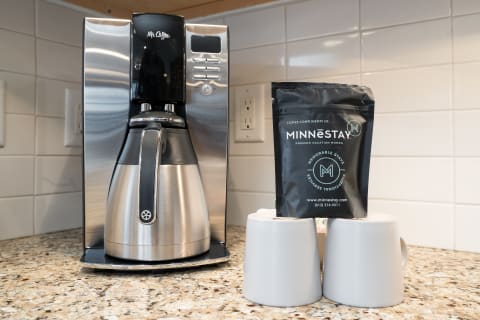 A modern coffee maker beside a package of premium coffee beans and two gray mugs on a granite countertop.