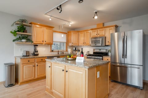 A spacious kitchen featuring light wood cabinets, granite countertops, and stainless steel appliances, with a central island and decorative plants.