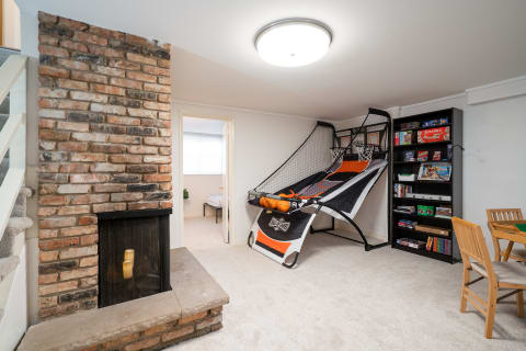 A game room featuring an arcade basketball setup, a brick fireplace, and a bookshelf full of games.