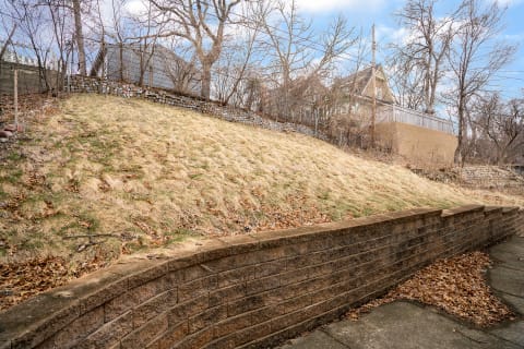 A sloping lawn with dry grass and trees, backed by a grey house, featuring a stone wall at the base.