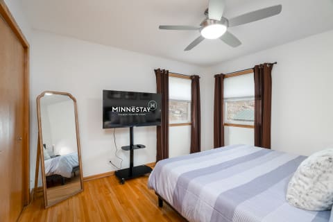 Bright bedroom with king-size bed, ceiling fan, and flat-screen TV on a stand, showcasing city views through large windows.