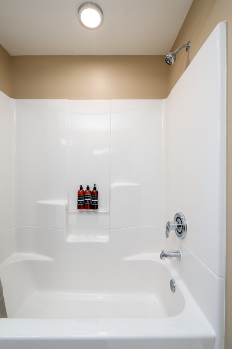 A modern tub-shower combo with white glossy surfaces and dark amber toiletries on a shelf.