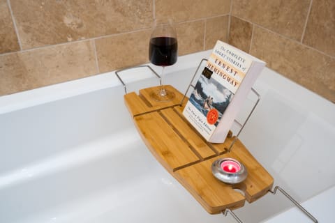 Bamboo bath caddy with a glass of wine, a book by Ernest Hemingway, and a candle, set on a bathtub for a relaxing atmosphere.