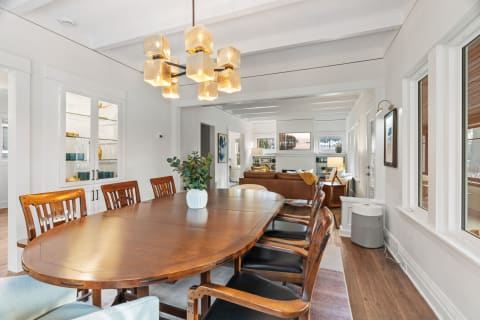 A dining room with a wooden table and modern chandelier, leading to a living area with a brown leather sofa.