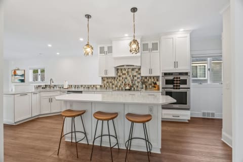 A modern kitchen with white cabinets, a spacious island, stylish pendant lights, and a warm wooden floor.