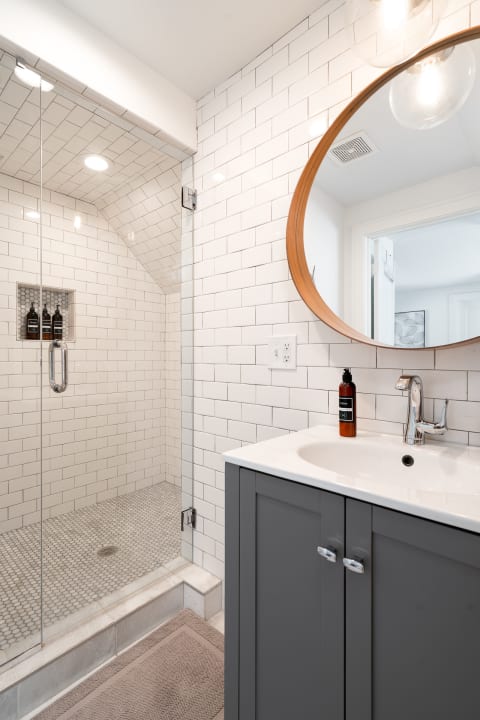 A contemporary bathroom with white subway tiled walls, a circular wooden mirror, and a glass shower enclosure.