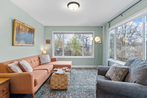 Stylish living room featuring an orange sectional couch, a woven ottoman, and large windows with a view.
