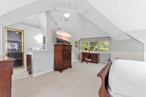 Bright attic bedroom featuring a sloped ceiling, wooden dresser with TV, and a workspace.