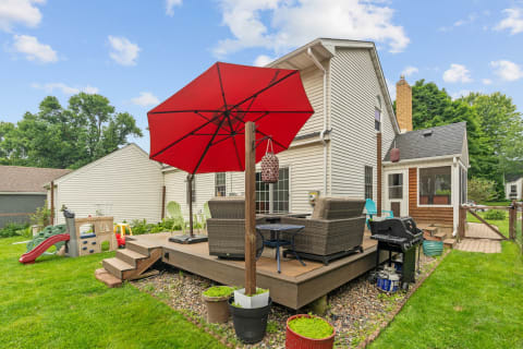 A backyard featuring a deck with seating under a red umbrella and a children’s play structure.