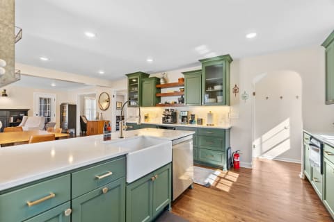 A cozy kitchen with green cabinets and a white countertop, featuring a farmhouse sink and an adjoining living space.
