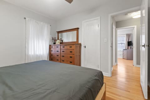 A bedroom with a grey bedspread, wooden dresser, and white curtains letting in natural light.