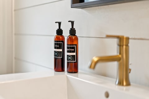 Amber bottles of hand soap and lotion sit elegantly on a white countertop near a gold faucet.