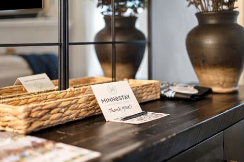A cozy welcome display on a wooden table with a woven basket, a thank you card, and a decorative vase of dried flowers.