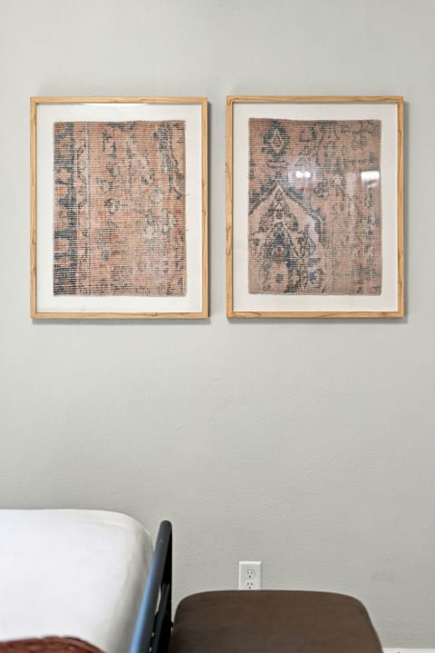 A minimalist bedroom wall with two framed prints displaying intricate patterns in warm colors, beside a bed and a bench.