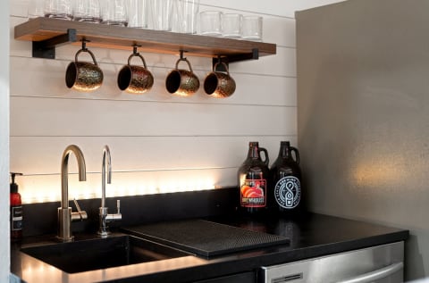 A cozy coffee bar featuring a rustic wooden shelf with mugs, a sleek dual-faucet sink, and decorative growlers.