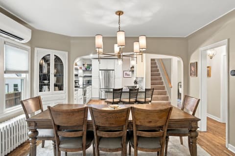 A cozy dining room with a wooden table, chairs, and a modern chandelier, adjoining a bright kitchen.