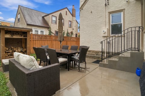 An outdoor patio with a dining table surrounded by wicker chairs and a cozy seating area, featuring a wood log storage.