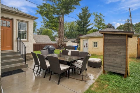 A patio with a dining table and chairs, an umbrella, a grill, and surrounding greenery.