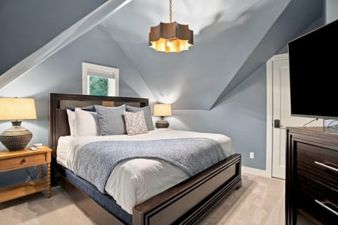 Cozy bedroom with vaulted ceilings, a wooden bed frame, decorative pillows, warm lighting, and a TV.