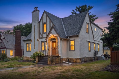 A Tudor-style home with warm lights glowing from its windows at dusk, surrounded by greenery.