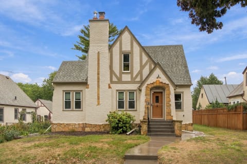 A beautiful Tudor-style house with textured stucco, stone accents, and a lush green lawn in a quiet residential area.