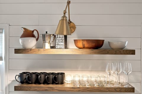 Wooden shelves with a pitcher, espresso maker, mugs, and glassware in a cozy kitchen.