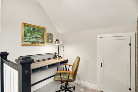 A cozy workspace under the eaves featuring a desk, vintage chair, framed artworks, and a lamp.