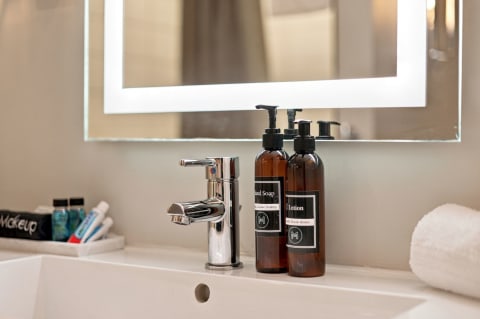 Modern bathroom sink with polished chrome faucet, amber bottles of hand soap and lotion, and a rolled white towel.