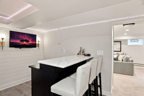 Stylish basement bar with white countertop, barstools, and a TV displaying a sunset scene.