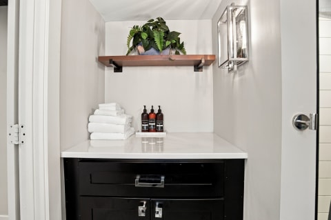 A modern bathroom corner with white towels and stylish toiletries on a reflective countertop, accented by a wooden shelf with plants.