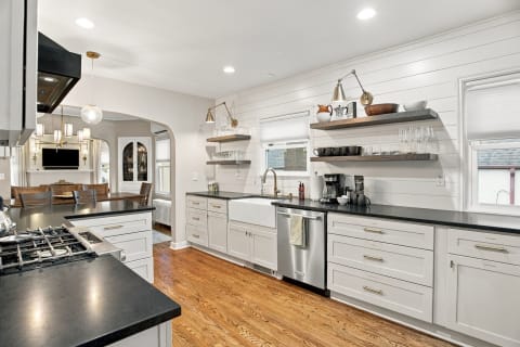 A spacious and modern kitchen with white cabinetry, dark countertops, and warm wood floors, featuring open shelving and a farmhouse sink.