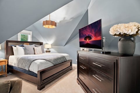 A cozy attic bedroom featuring a king-sized bed, a dresser with a TV, soft lighting, and decorative flowers.
