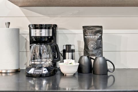 A coffee maker, a bag of Minnestay coffee, and two black mugs arranged on a kitchen countertop.