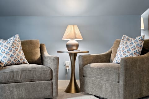 Cozy seating area with two gray armchairs, decorative pillows, and a stylish lamp on a bronze table against blue walls.