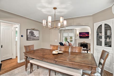 A cozy dining room featuring a wooden table, plush benches, and a modern chandelier, with an open view to a living area.