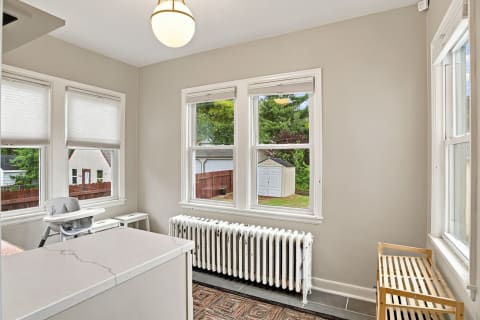 A bright nursery with large windows, a white radiator, and a wooden bench, overlooking a serene garden.