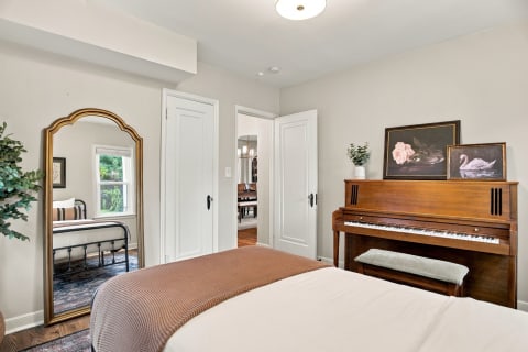 A cozy bedroom with white linens, a vintage piano, and an ornate mirror reflecting the space.