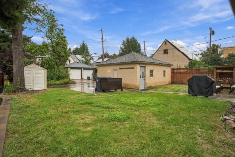 A spacious backyard with a grassy area, utility shed, and garages against a blue sky.