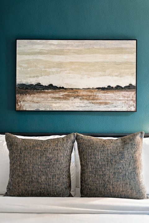 A framed landscape painting above a white bed with checked pillows against a teal wall.