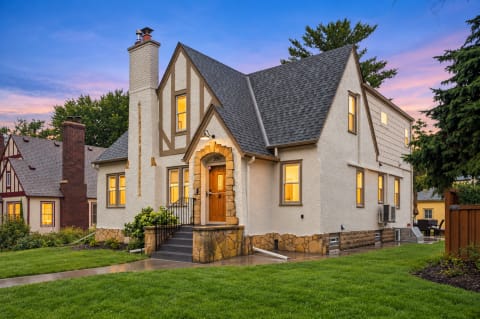 Tudor-style home with peaked roofs, stone chimney, and warm window lights surrounded by greenery and twilight sky.
