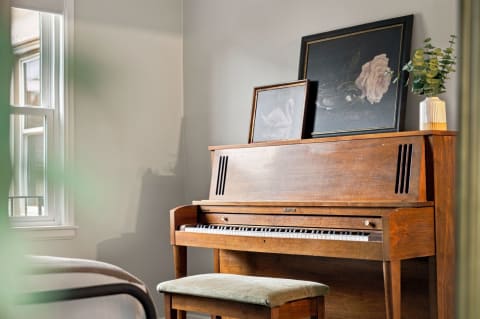A vintage piano with framed art and a vase of eucalyptus stands in a sunlit room.