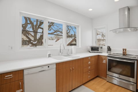 Modern kitchen with white countertops and wood cabinetry, featuring large windows overlooking a tree.