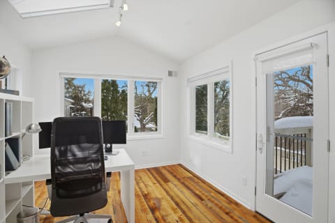 A modern home office with a white desk, black chair, and large windows showcasing snowy trees outside.